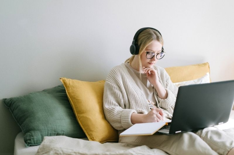 Caucasian girl in sweater with headphones sitting on sofa with laptop on her thighs